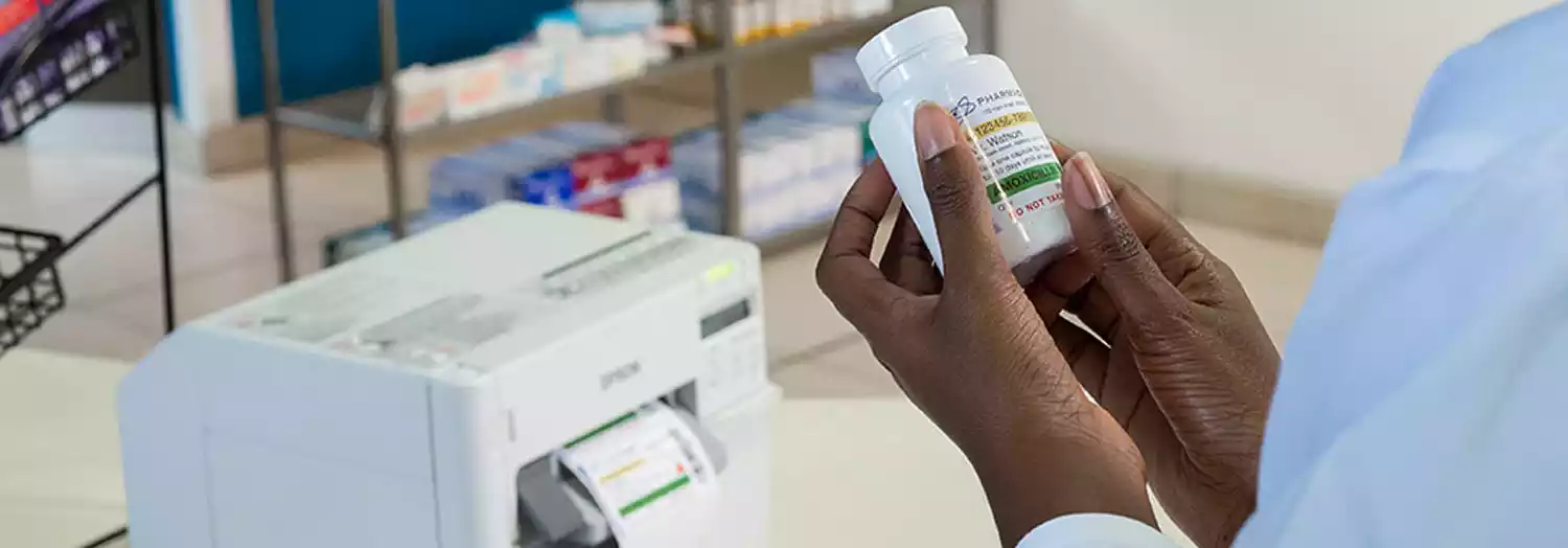 Manos sosteniendo frasco de medicamento etiquetado y al fondo, la etiquetadora EPSON ColorWorks C3500 (TM-C3500)
